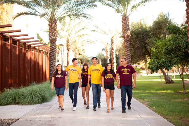 A group of six individuals wearing maroon and gold t-shirts with ‘SUNDEVIL’ printed on them walk side by side on a pathway lined with tall, narrow trees. The text ‘Fall 2024’ is displayed above them.