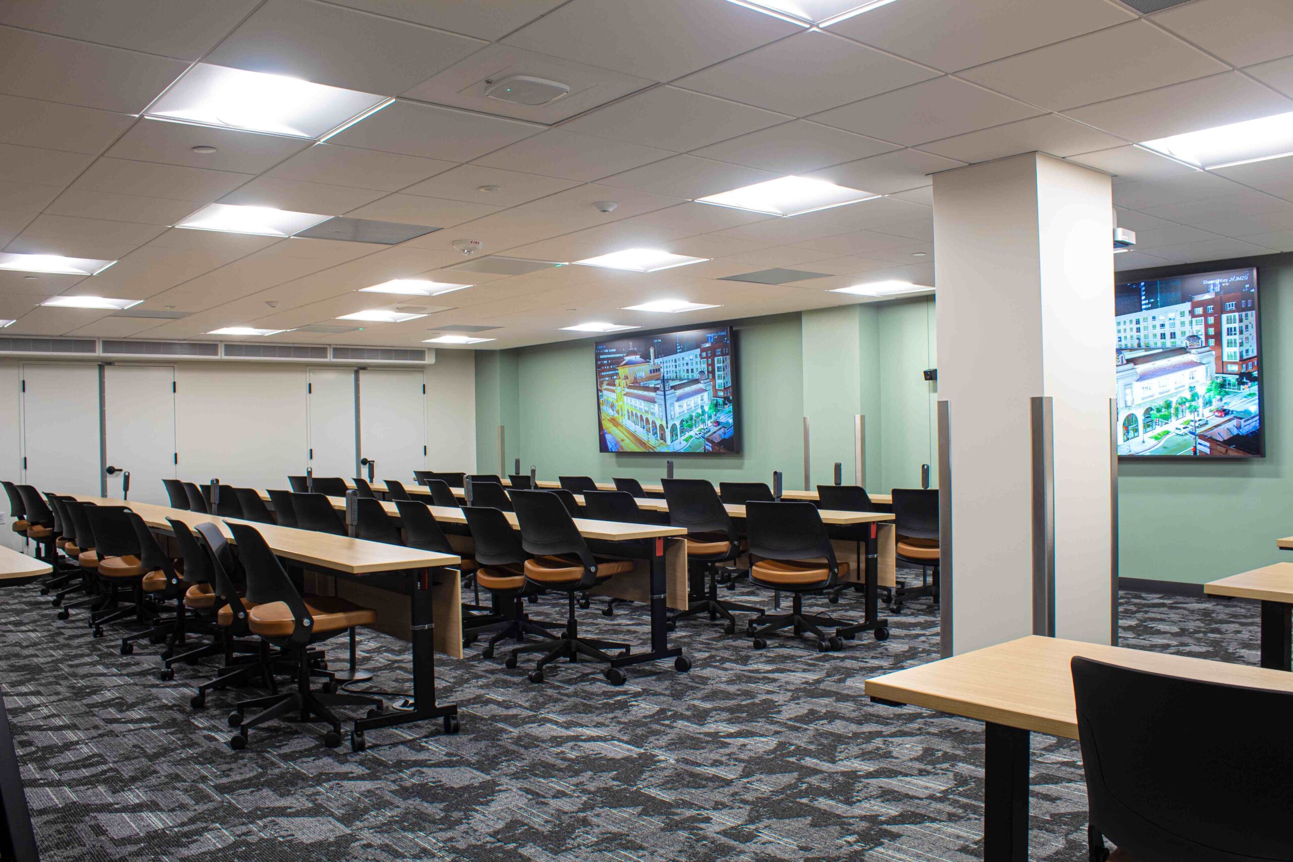 A modern classroom with rows of long tables and ergonomic chairs, arranged in a lecture-style setup. The room features a carpeted floor, ceiling lights, and multiple large screens mounted on the walls displaying images of buildings. The walls are painted in a light green color, and the space is well-lit, creating a clean and organized learning environment.