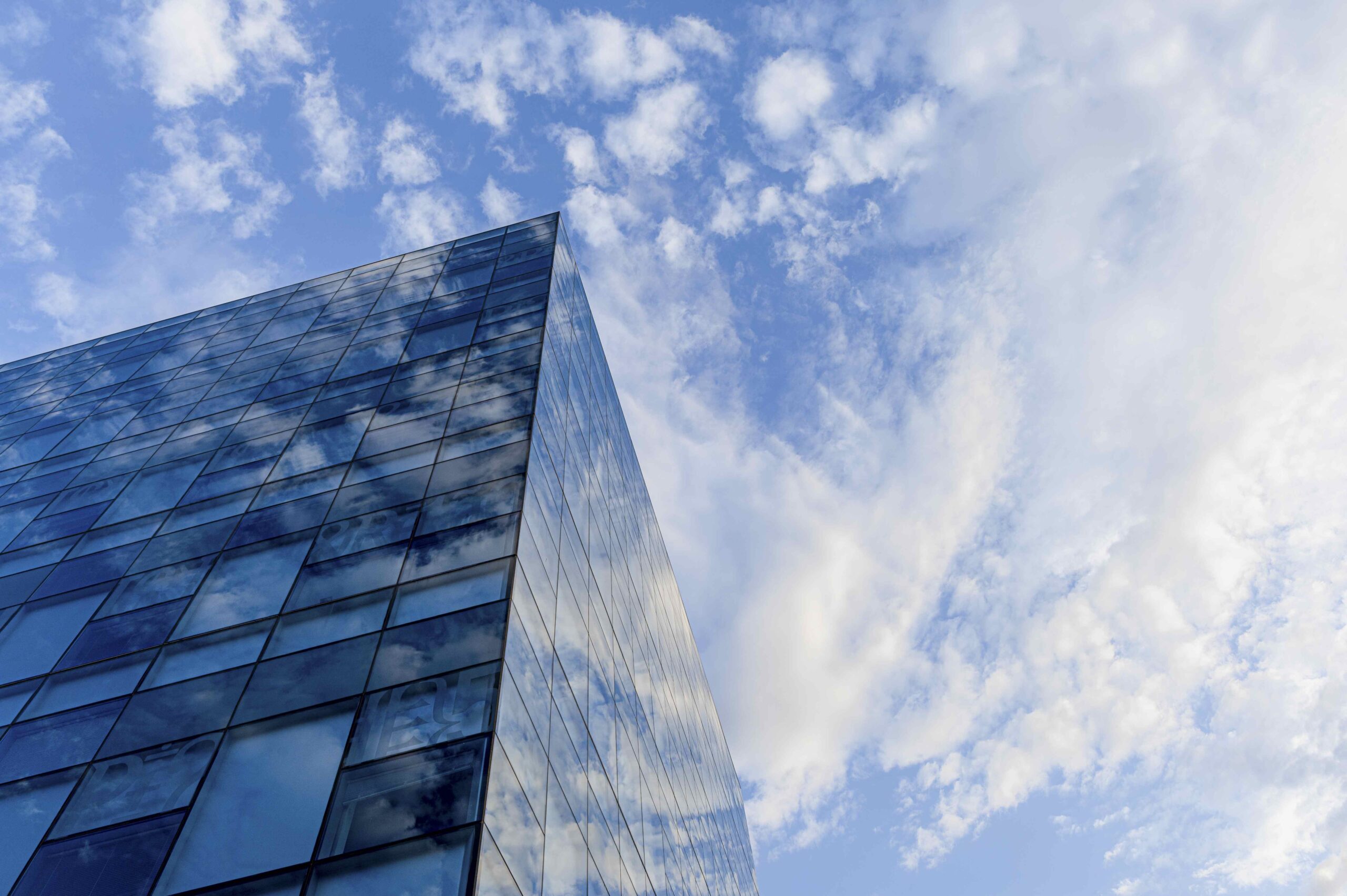 A modern glass building with reflective windows, capturing the blue sky and clouds. The perspective shows the building's sharp corner extending upwards into the sky, emphasizing the clean lines and contemporary architecture. The sky is partly cloudy, with soft white clouds scattered across the blue backdrop, creating a serene and open atmosphere.