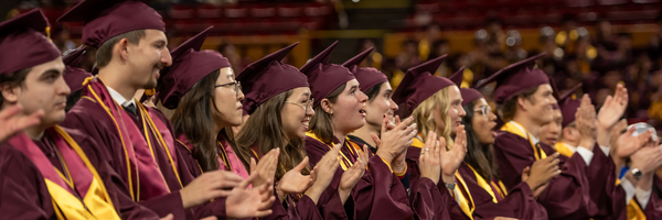 Group of graduates in maroon caps and gowns, clapping and celebrating at a commencement ceremony.