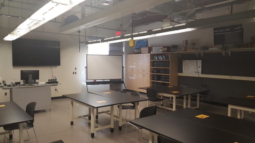 Photograph of a classroom (PRLTA 103). Shown are many desks. On the far wall is a large screen. The instructor's workstation is located in front of the screen. A white board and a bookrack is located along the adjacent wall.