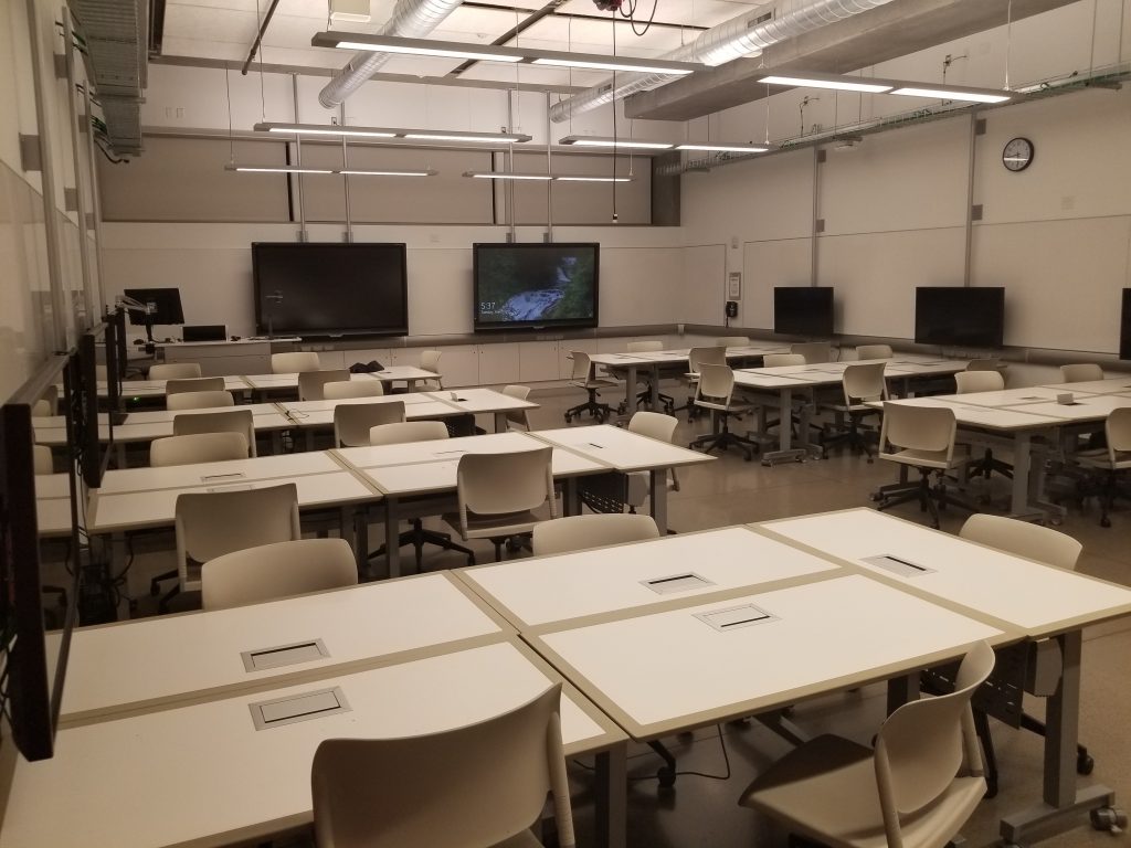 A classroom with several desks, chairs and TV screens.