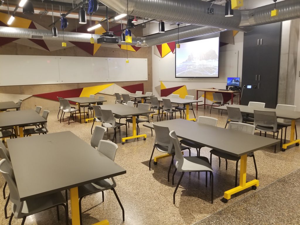 Photograph of a classroom (TKDR D101). Shown are many tables and chairs . On the far wall is a large projector screen. In front of the screen the instructor's work station is visible.