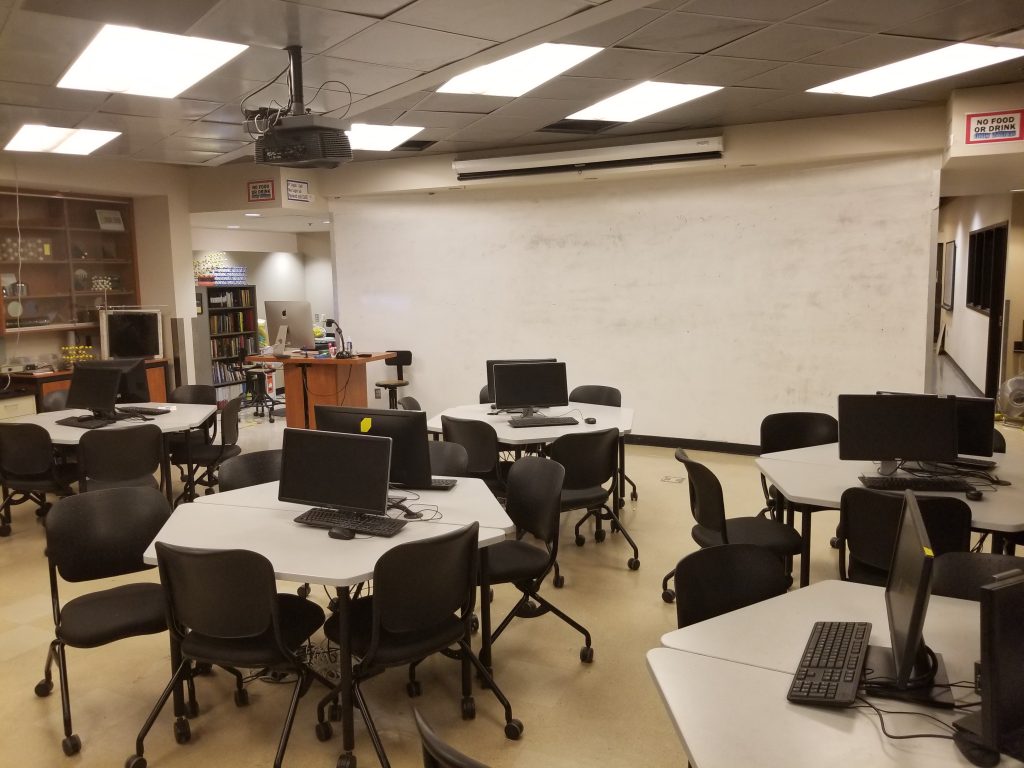 A lab that has several round tables with monitors and a whiteboard at the back.
