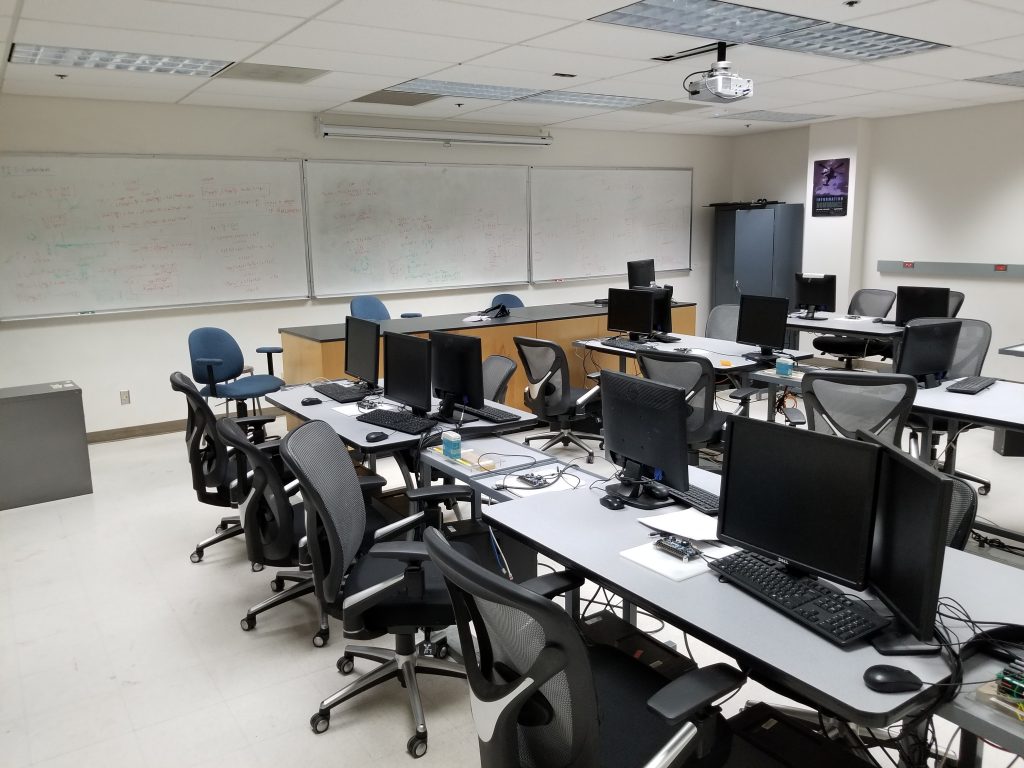 A computer lab equipped with  several desks, chairs, monitors, an instructor table and white boards.