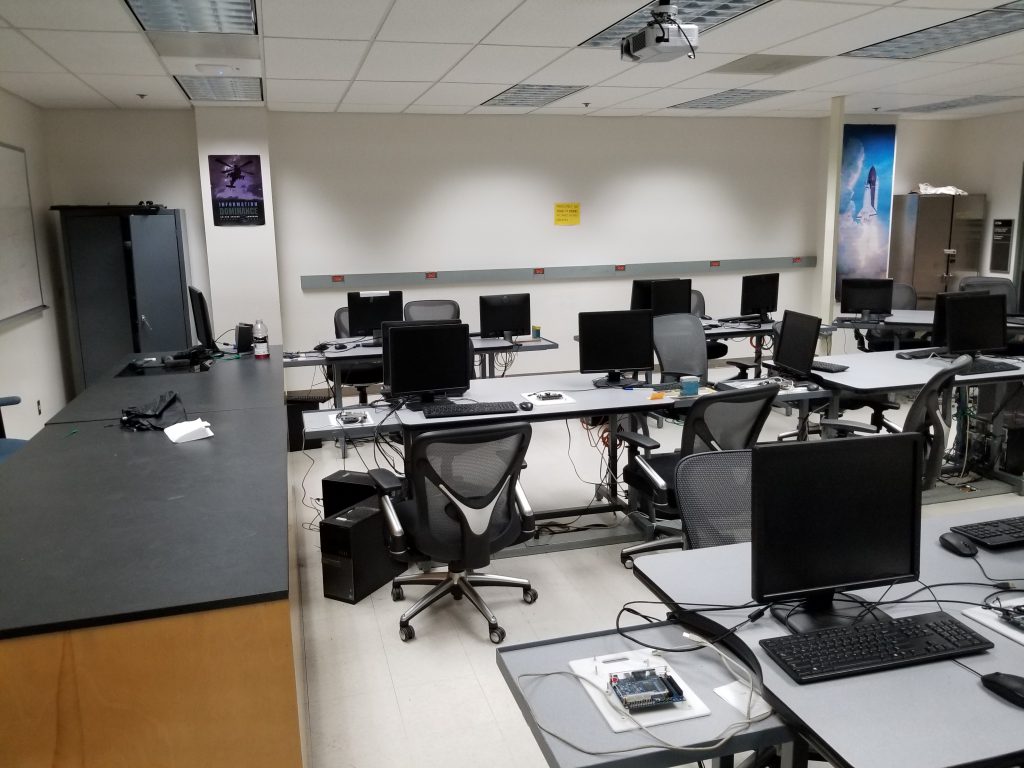 A computer lab equipped with  several desks, chairs, monitors, and an instructor table.