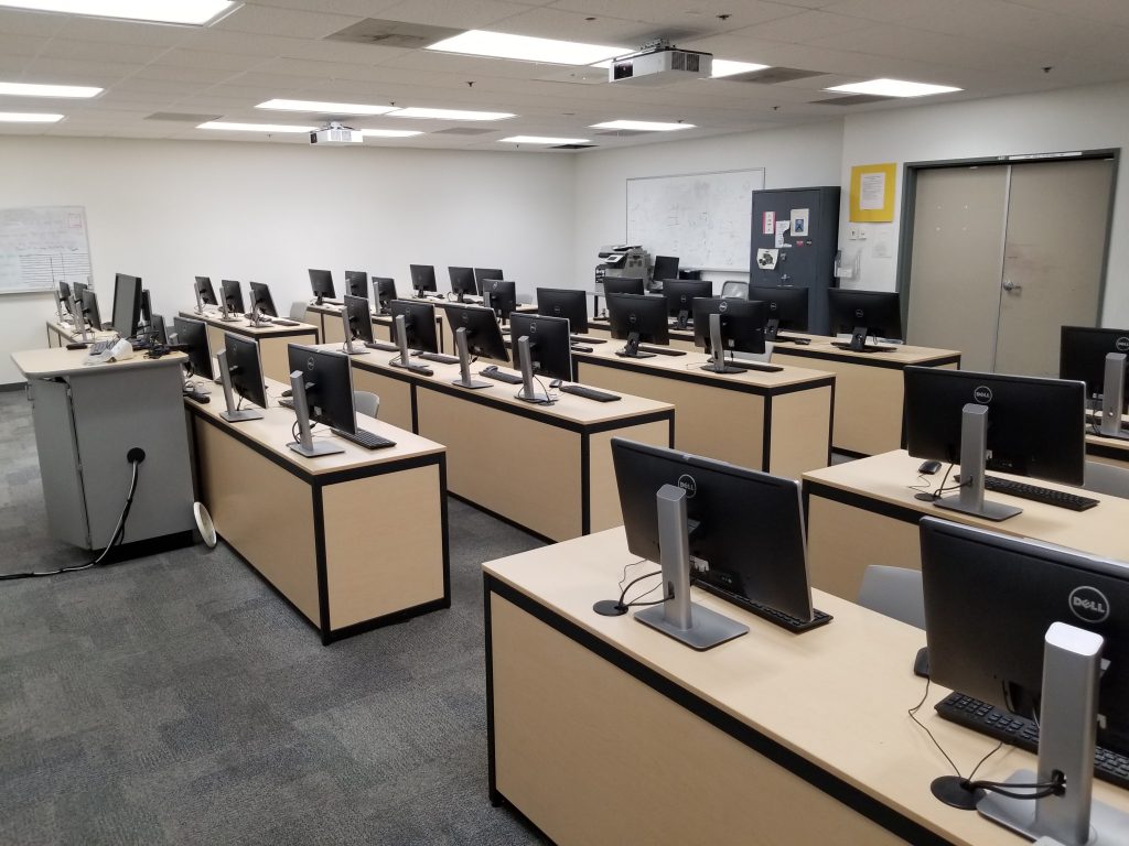 An instructor lab with several monitors, desks, keyboards, mouse and projector screens.