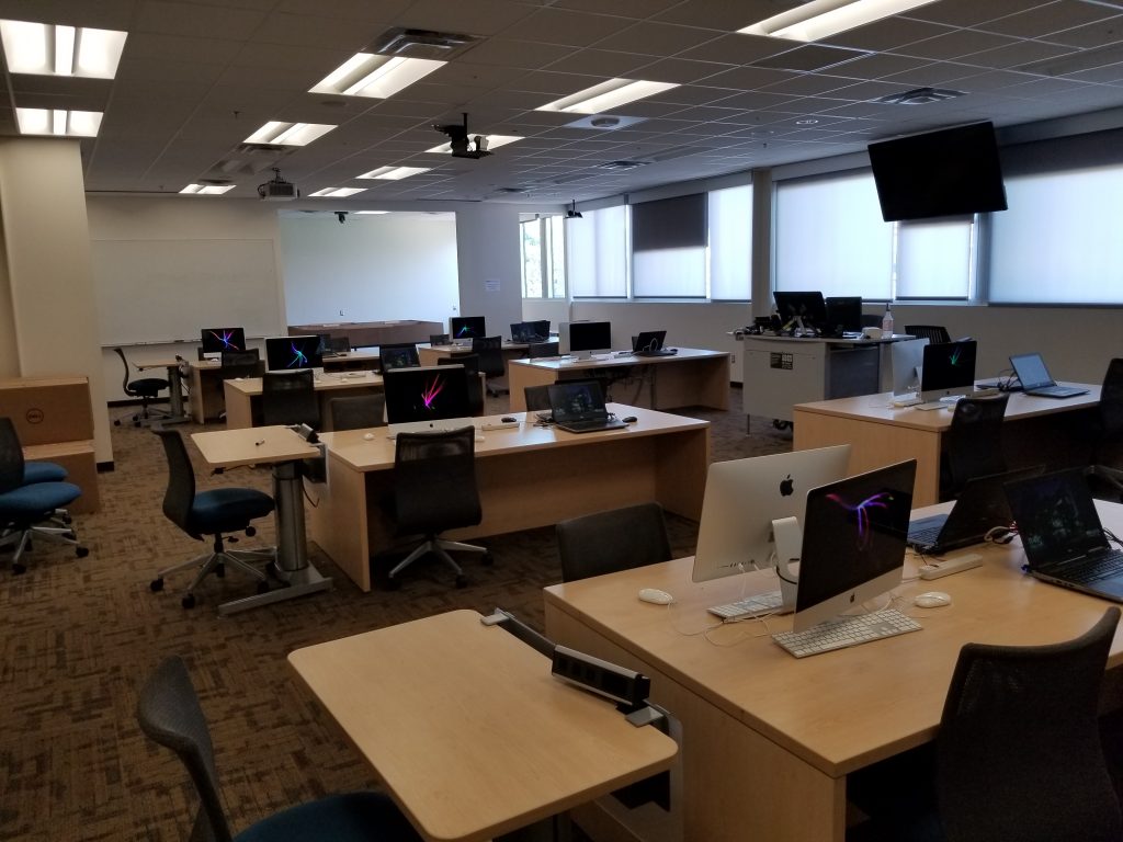 A lab with several desks, each equipped with two mac monitors, two laptops, and chairs. There is also a workstation for instructors. On the far wall is a large white board. 