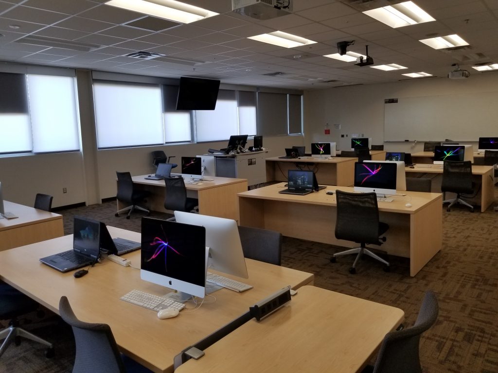 A lab with several desks, each equipped with two mac monitors, two laptops, and chairs. There is also a workstation for instructors. On the far wall is a large white board. 