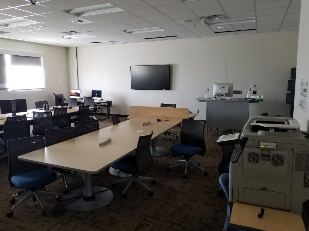 A classroom with 2 vertical rows of tables and chairs, a TV Screen, an instructor station and a printer.