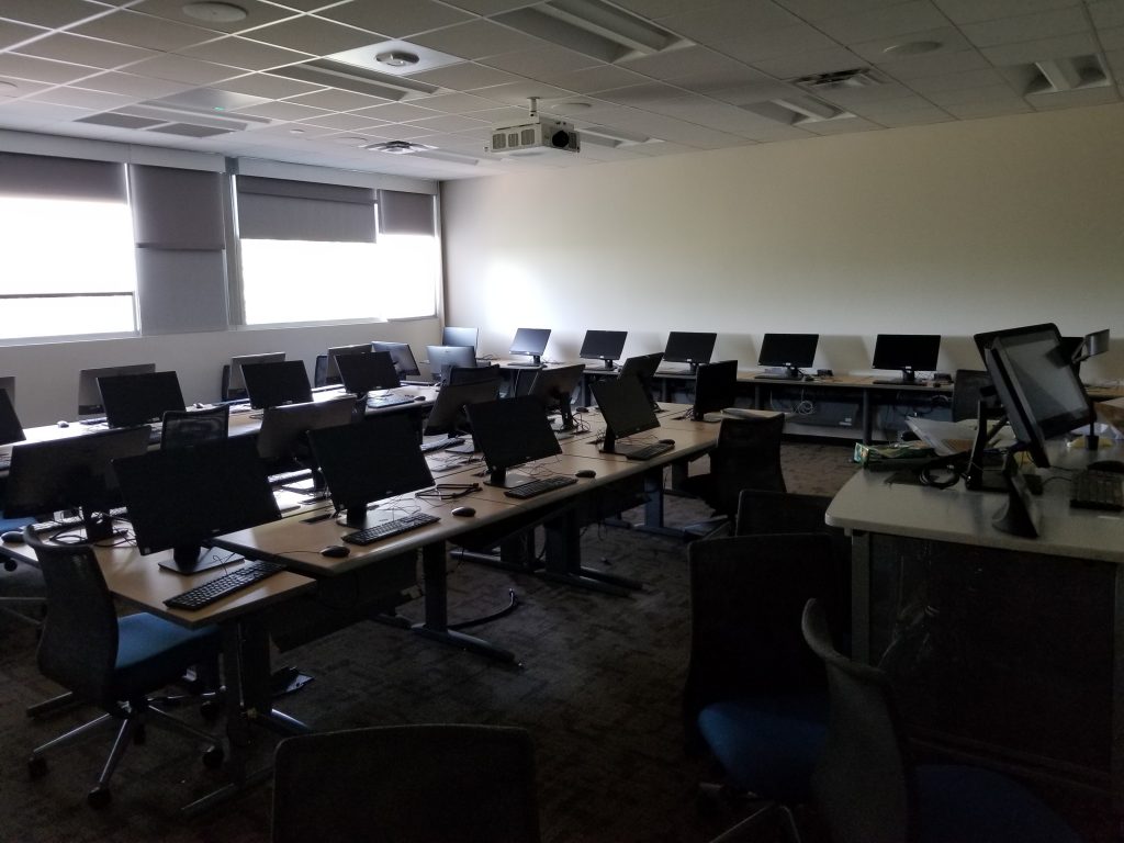 A lab with several rows of desks and monitors, few chairs, a projector and an instructor station.