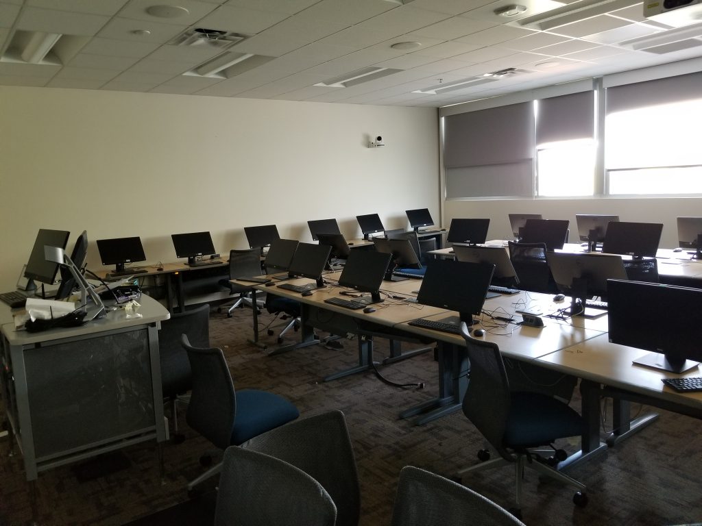 A lab with several rows of desks and monitors, few chairs and an instructor station.