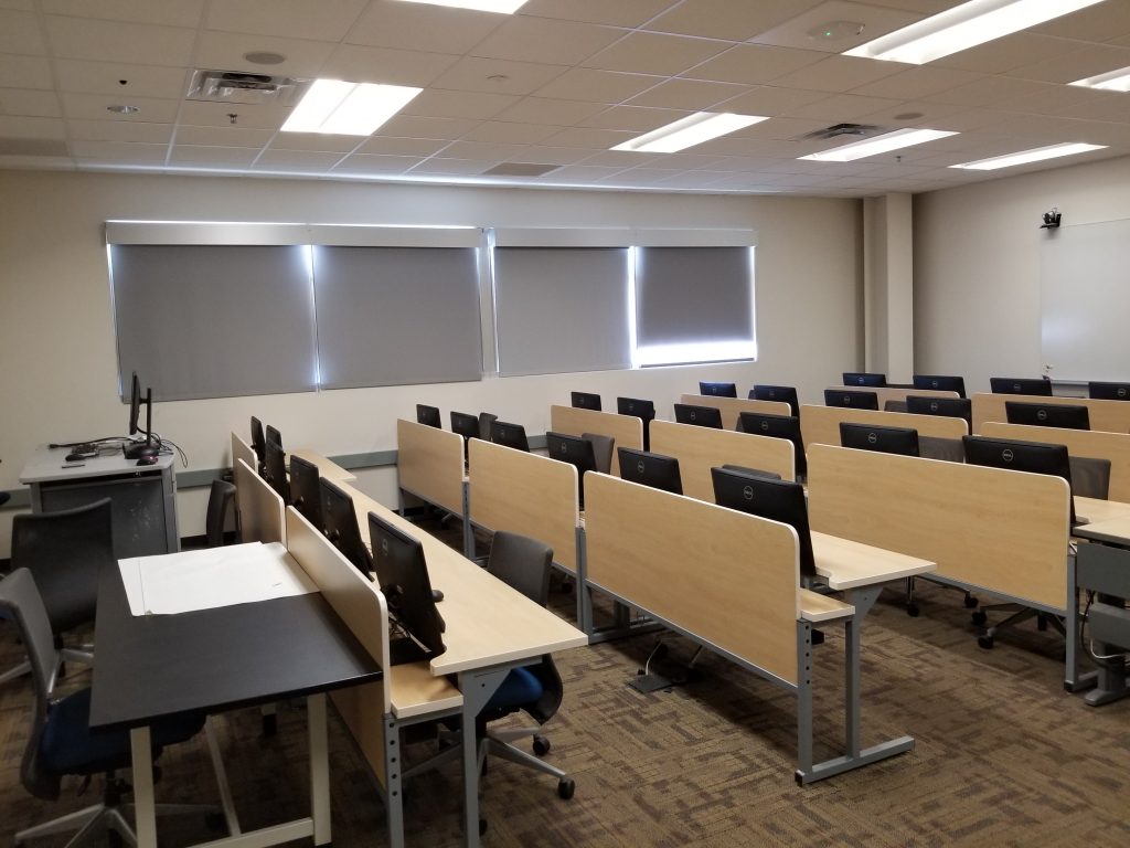 Photograph of a classroom (PRLTA 202). Shown are many desks, each with two computers (the backs of which are visible.). There is also an instructor's station, which has a monitor. 