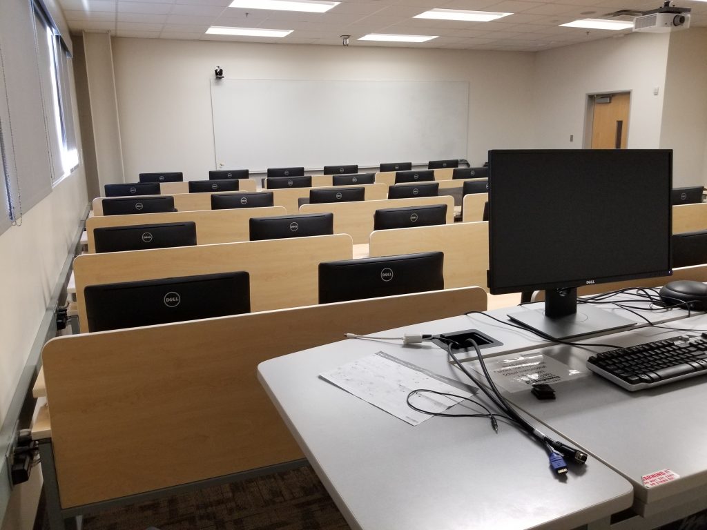 Photograph of a classroom (PRLTA 202). Shown are many desks, each with two computers (the backs of which are visible.). There is also an instructor's station, which has a monitor. The far wall has a large whiteboard.