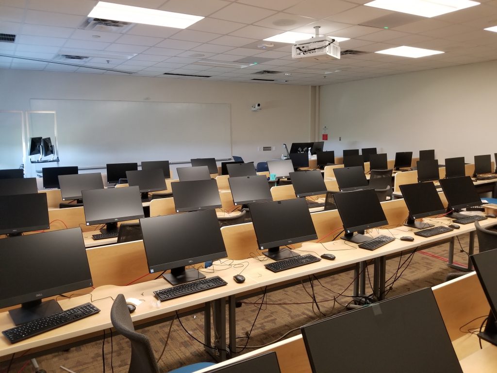 Photograph of a classroom (PRLTA 201). Shown are many desks, chairs and computers (the screens of which are visible). On the far wall is a large whiteboard.  front of the whiteboard the instructor's work station is located.