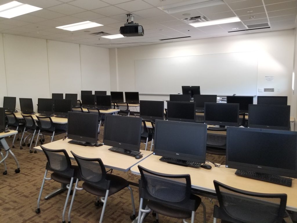 Photograph of a classroom (SANCA 153). Shown are many desks, each with two chairs and two computers (the screens of which are visible). On the far wall is a large whiteboard. Above the white board are two projector screens, which are retracted. In front of the whiteboard the instructor's work station is visible.