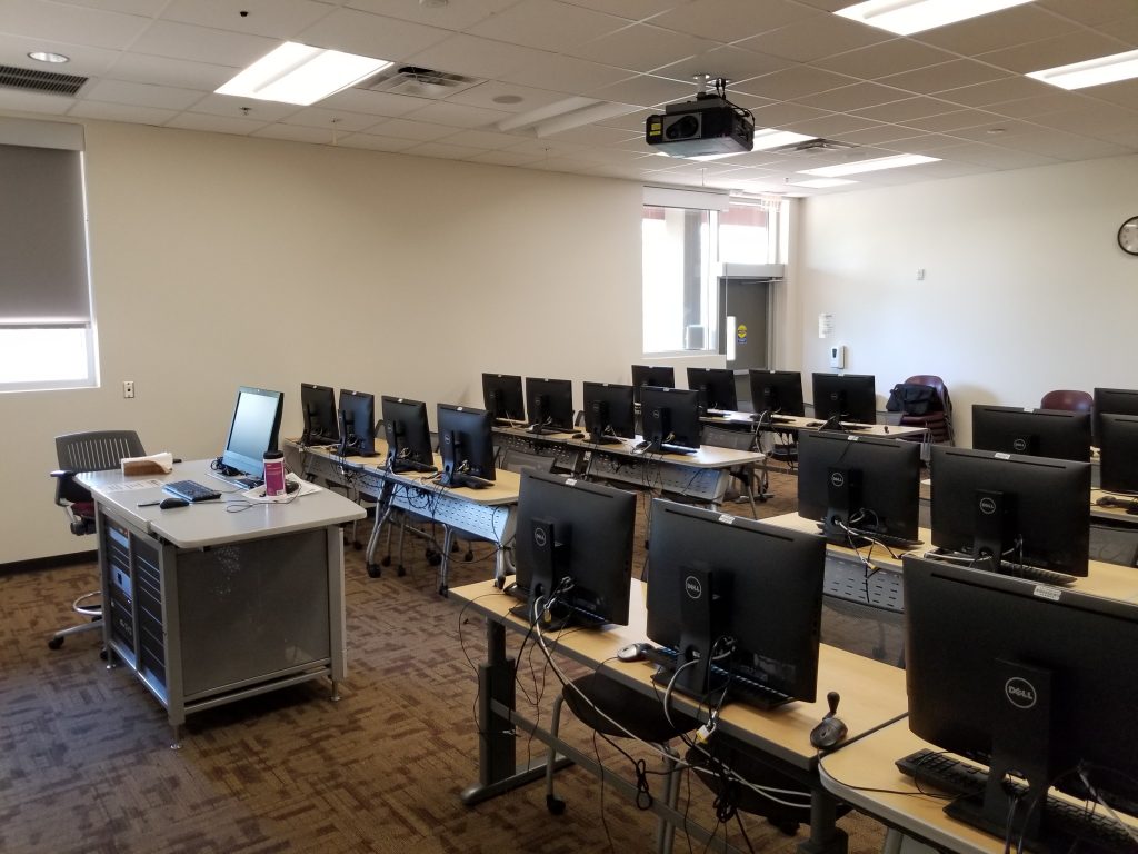 Photograph of a classroom (SANCA 153). Shown mounted on the ceiling is a projector. To the left is the instructor's work station: a desk with a computer, in which the screen is visible, and other equipment. Next to it a chair. The rest of the room is filled with desks, each desk has two computers, the backs of which are visible.