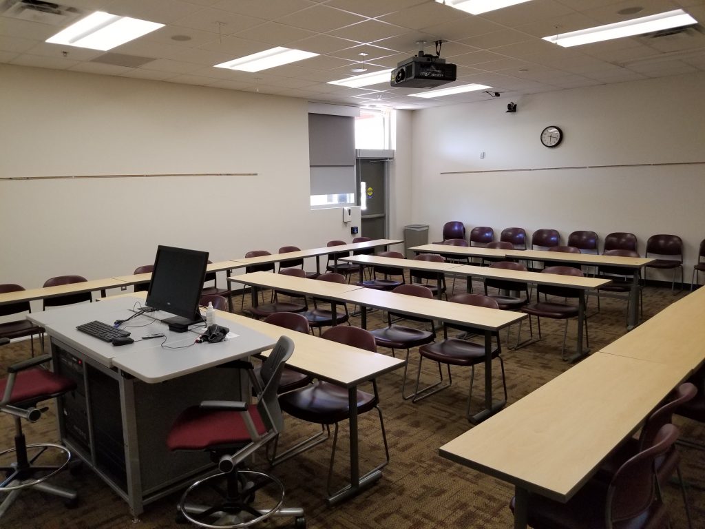 Photograph of a classroom (SANCA 151). Shown are many desks. In the classroom, there is a workstation for the instructor with a monitor.