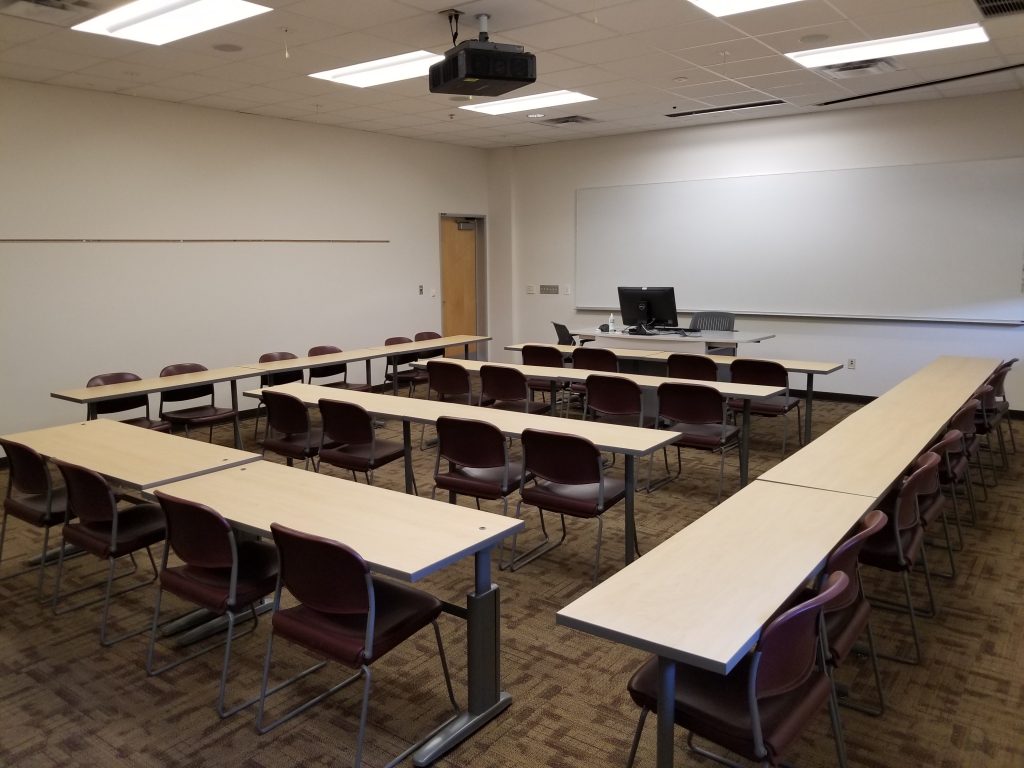 Photograph of a classroom (SANCA 151). Shown are many desks. On the far wall is a large whiteboard.  An instructor's workstation with a monitor is present in the classroom.