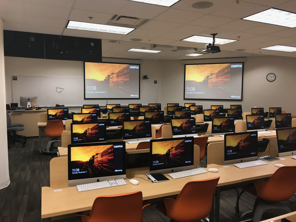 A lab that has several rows of mac monitors, an instructor station and a couple of projectors.