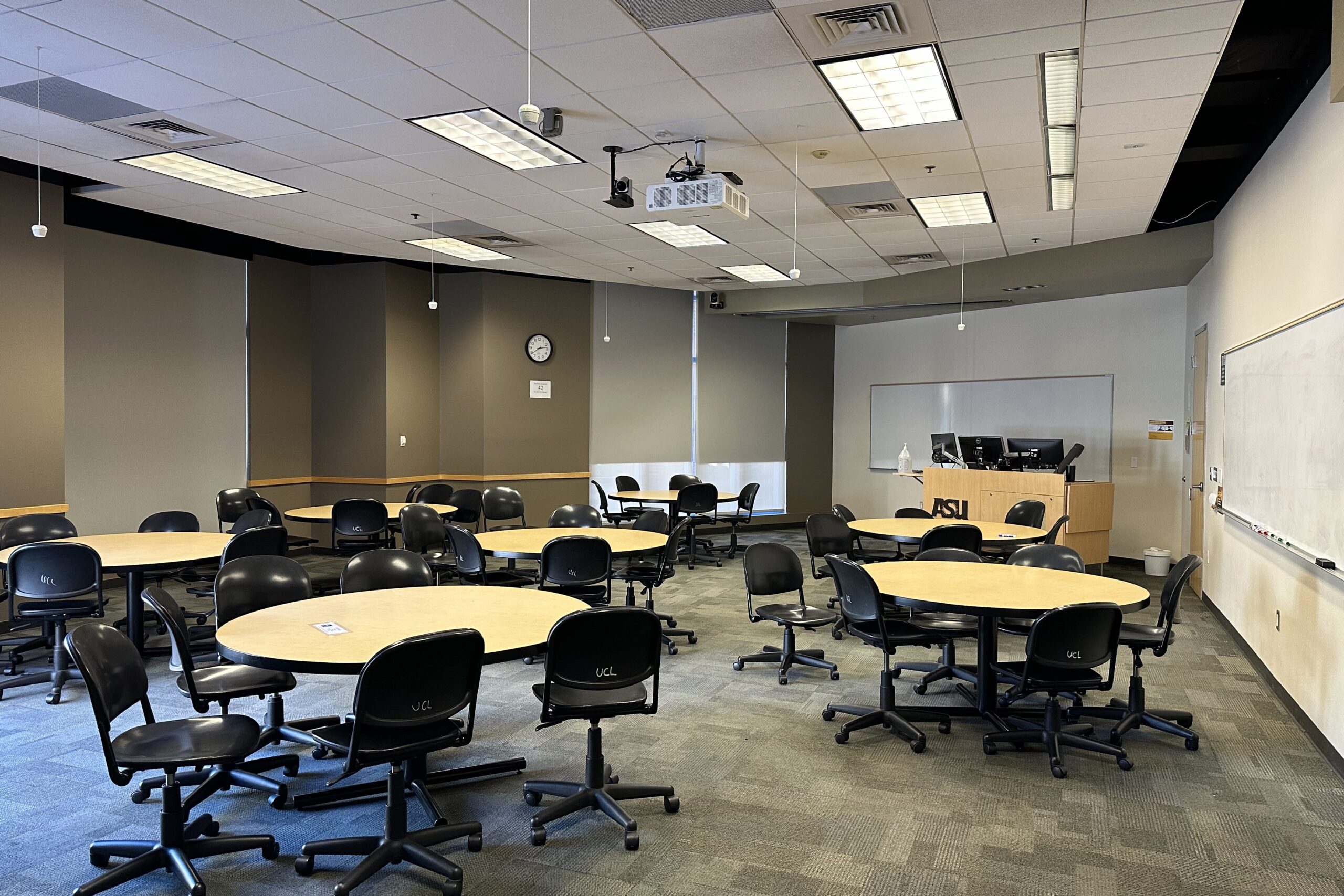 A classroom in the Brickyard building. Multiple round tables with chairs surrounding them. An instructor work station and two white boards, one behind it and one to the side.