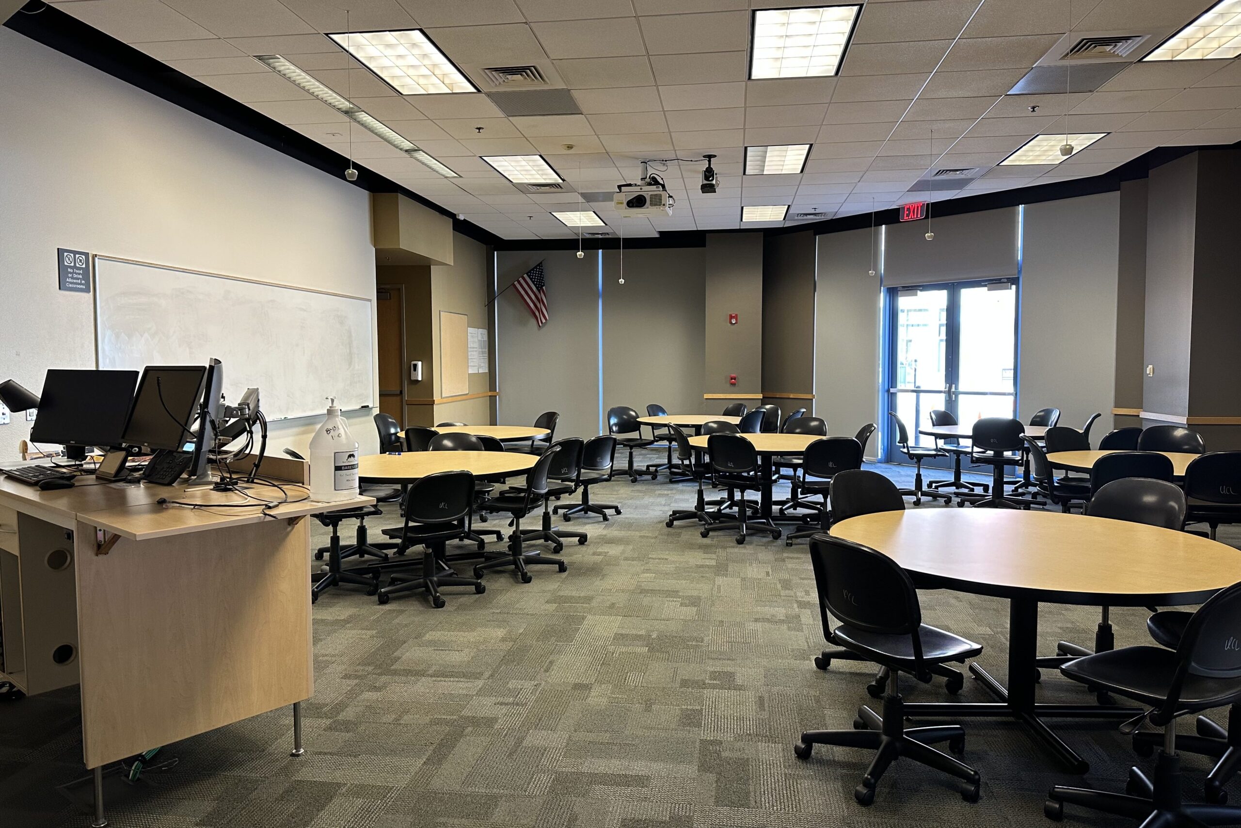 Classroom in the Brickyard building. There are seven round tables with chairs surrounding, a white board, projector and doors on the left that go into the building and to the right that exit the building.