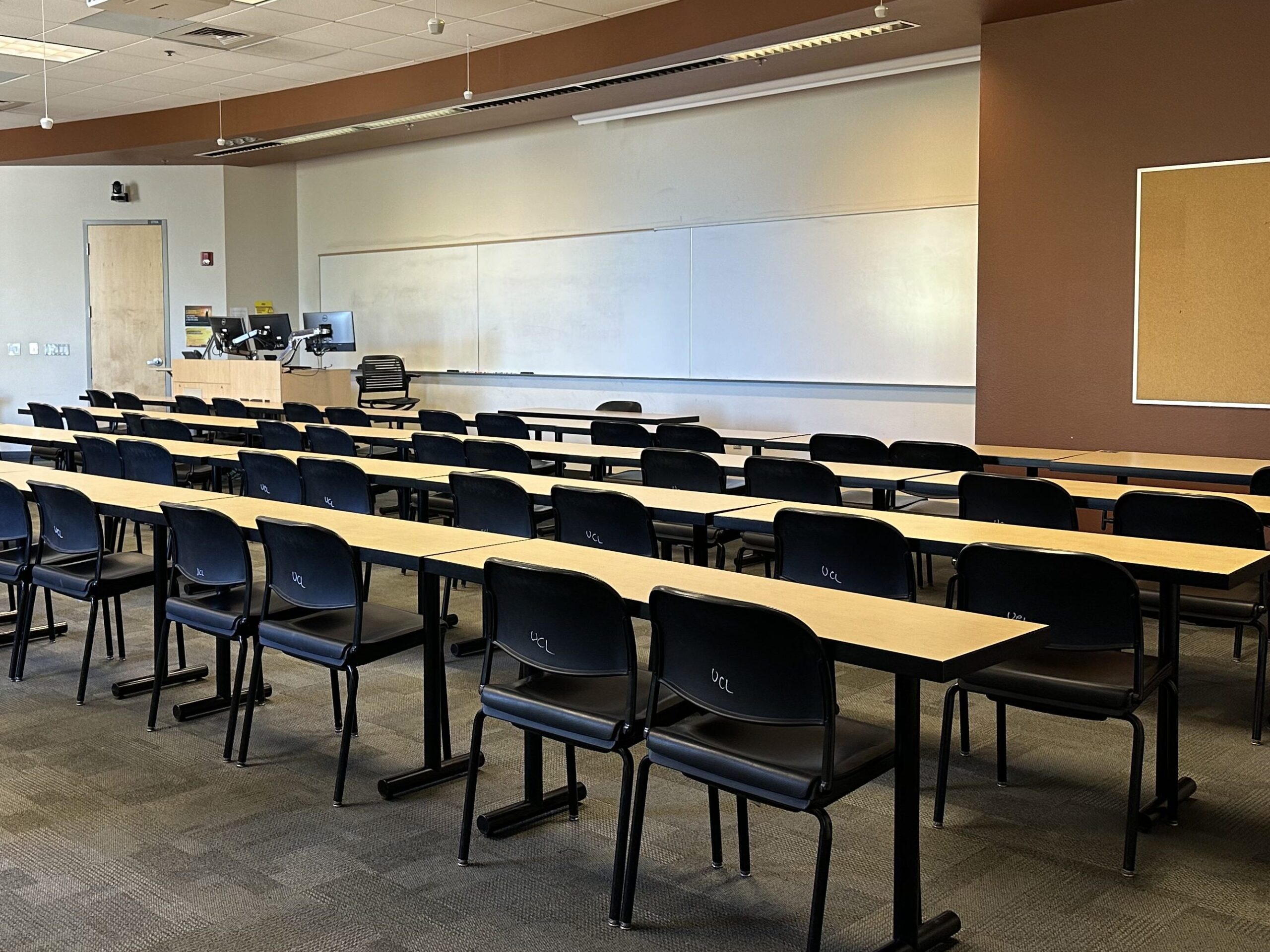 Classroom in ASU's Brickyard building. Rows of tables, lined with chairs, all facing the front of the room. The wall has a long whiteboard and instructor podium. There is a door to the left with a camera above it. From the ceiling hang microphones.