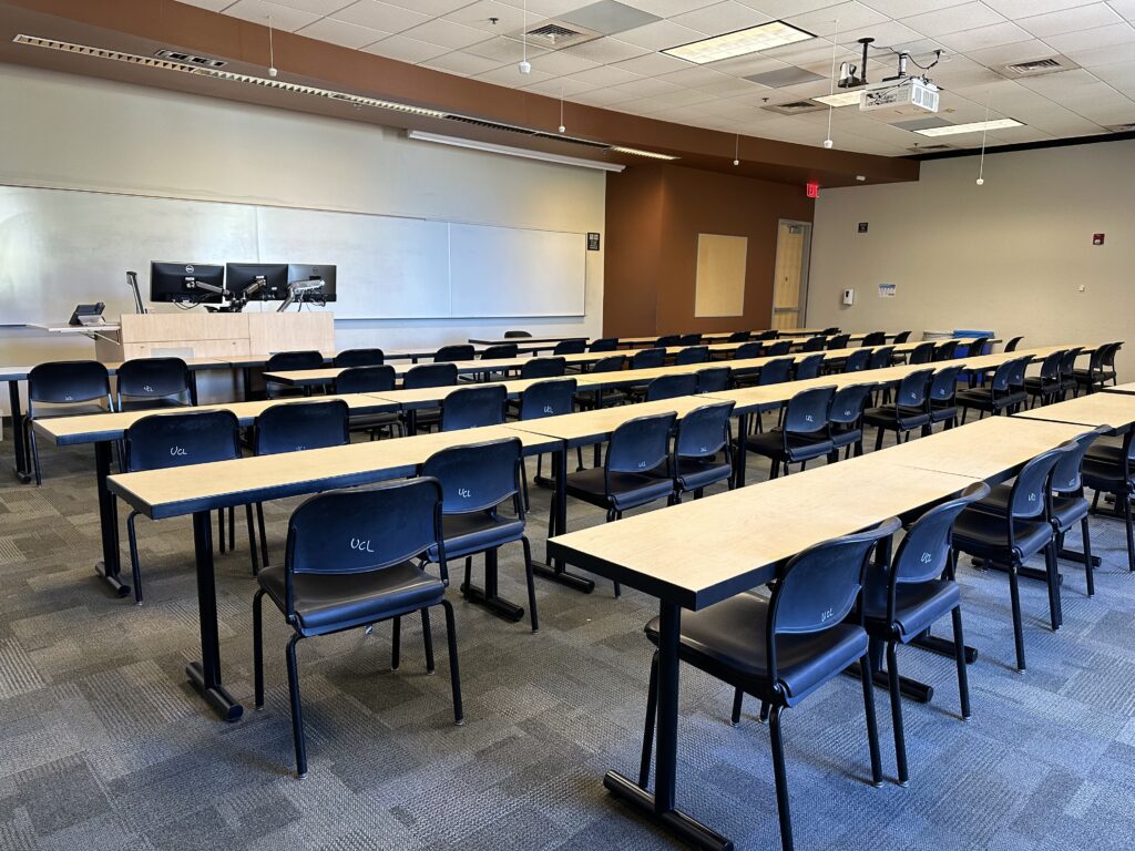 A classroom in the Brickyard building. Long tables lined with chairs are facing the front of the room. Here you see the instructor podium and behind it a long whiteboard. The ceiling is adorned with speakers and a projector. To the right is a door to exit the room.