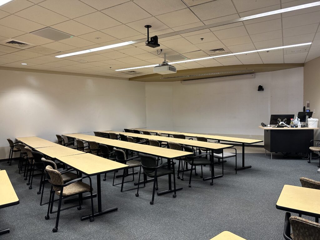 A classroom belonging to the Ira A. Fulton Schools of Engineering. Shown is the backside of the desks and the projector, a class camera on the far wall, instructor work station and the screen used by the projector.