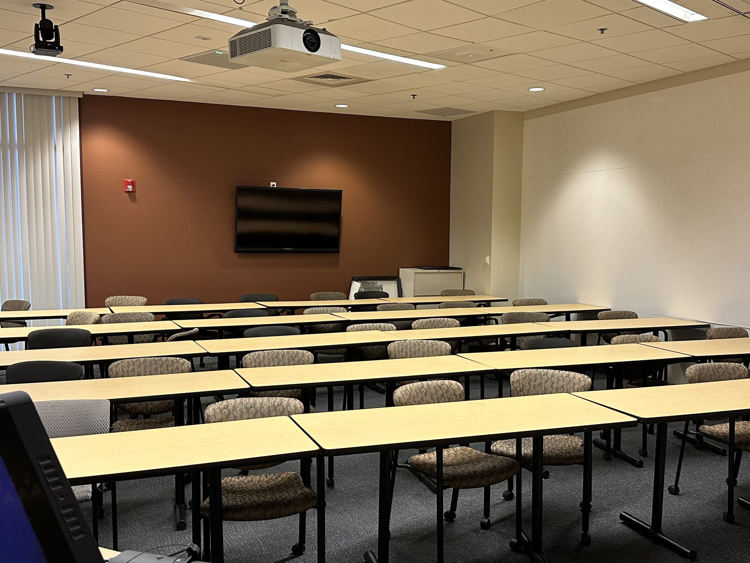 Classroom belonging to the Ira A. Fulton Schools of Engineering. Shown are many desks and chairs and a projector, a monitor on the back wall and ceiling microphone.