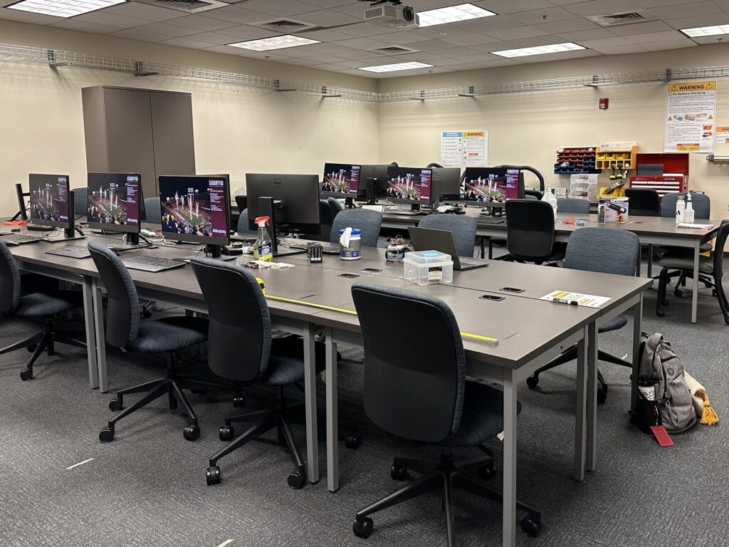 Computer lab with multiple windows workstations: desks with computers and rolling chairs. There is shelving on the wall, a short distance from the ceiling. In the back is a table with equipment and cubbies.