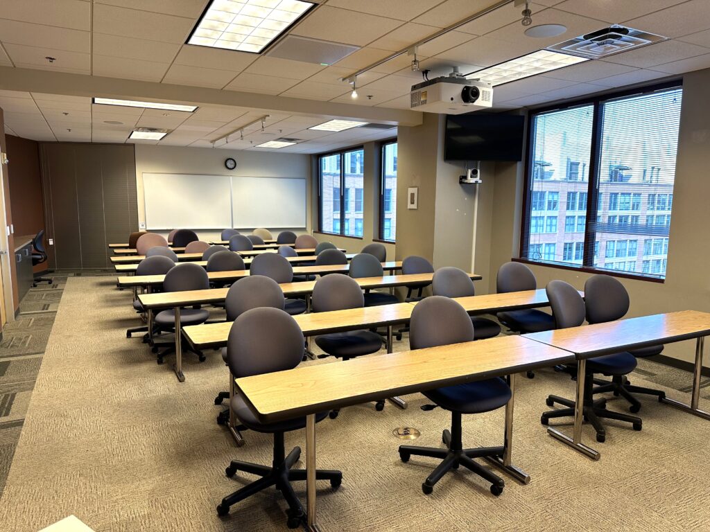 Classroom with large windows on the far wall, a projector on the ceiling, a TV on the wall with a camera under it. Whiteboards are on the back wall. There are rows of tables lined with chairs facing toward the camera.